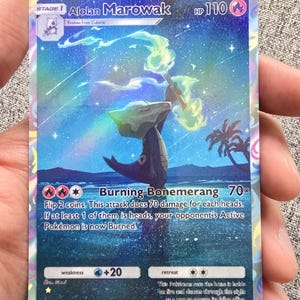May include: A holographic Pokémon trading card featuring Alolan Marowak. The card displays a colorful illustration of the Pokémon in a tropical setting with text that reads "Burning Bonemerang 70x". The card is held in a hand.
