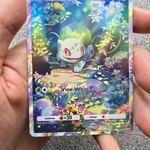 May include: A Pokemon trading card featuring Bulbasaur, a grass-type Pokemon. The card is holographic and has a light blue, green, and purple background with a floral pattern. The card has a green border and the text "BASIC Bulbasaur HP70" at the top. The card also has a picture of Bulbasaur in the center, and the text "Vine Whip 40" at the bottom.