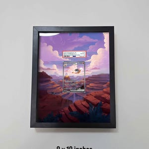 May include: A framed Pokemon card, 8 x 10 inches, featuring a Charizard illustration against a scenic background of a canyon and a sunset sky. The black frame is mounted on a white wall.