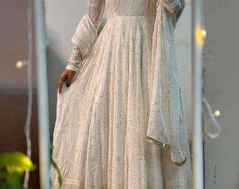 May include: A white, floor-length dress with intricate embroidery and a matching shawl. The dress features a square neckline, long sleeves, and delicate detailing. The shawl has a gold trim. The dress is reflected in a mirror.