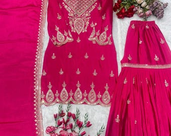 Può includere: Un vivace completo in tre pezzi color fucsia. Il set comprende una tunica a maniche lunghe con ricami dorati intricati, una gonna abbinata e una sciarpa. La tunica e la gonna presentano decorazioni dettagliate. L'outfit è esposto su una superficie bianca con accenti floreali.