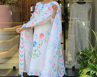 May include: A white floral print salwar kameez ensemble. The outfit includes a long-sleeved tunic, matching pants, and a sheer dupatta. The floral pattern features pink, blue, and yellow flowers. The dupatta has a chevron design on the border.