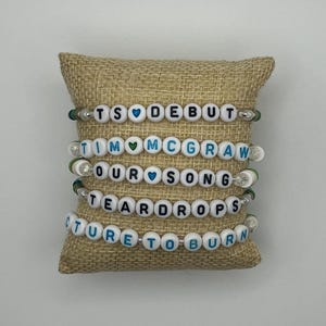 May include: Five beaded bracelets with white letter beads spelling out song titles, including "TS DEBUT", "TIM MCGRAW", "OUR SONG", "TEARDROPS", and "PICTURE TO BURN". The bracelets are displayed on a tan burlap pillow.