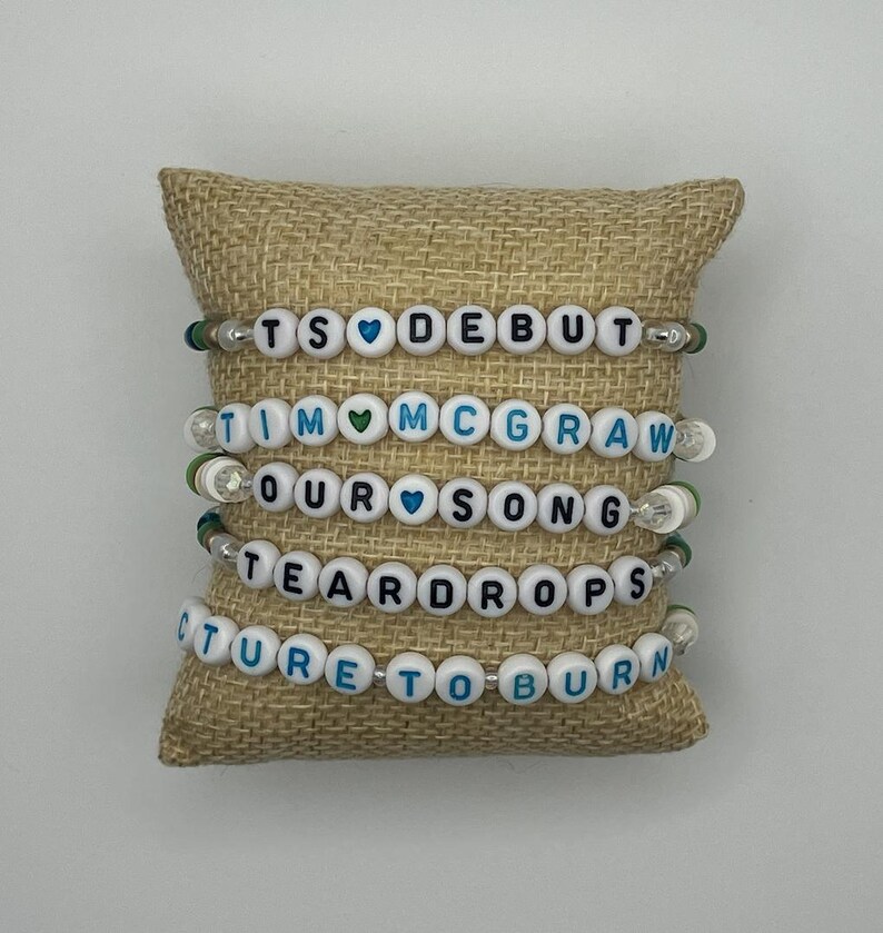May include: Five beaded bracelets with white letter beads spelling out song titles, including "TS DEBUT", "TIM MCGRAW", "OUR SONG", "TEARDROPS", and "PICTURE TO BURN". The bracelets are displayed on a tan burlap pillow.