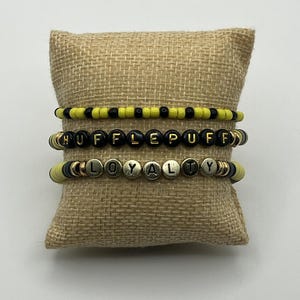以下が含まれることがあります： 黄、黒、金のビーズブレスレット3本がジュートのクッションに飾られています。一番上のブレスレットは黄色と黒のビーズです。真ん中のブレスレットは黒い文字で「HUFFLEPUFF」と綴られています。一番下のブレスレットは「LOYALTY」と綴られています。