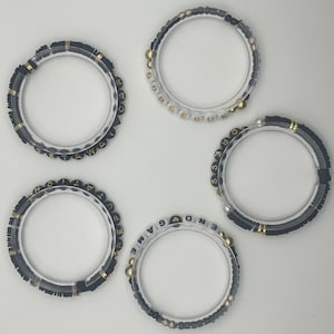 May include: Five clear acrylic bracelets with black and gold beads. Each bracelet has a different word spelled out in gold and black letter beads: "GETAWAY", "GORGEOUS", "DRESS", "REPUTATION", and "END GAME".