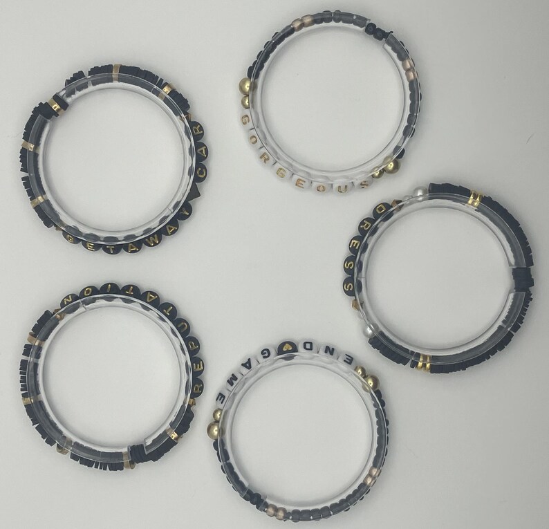 May include: Five clear acrylic bracelets with black and gold beads. Each bracelet has a different word spelled out in gold and black letter beads: "GETAWAY", "GORGEOUS", "DRESS", "REPUTATION", and "END GAME".