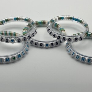 May include: Five clear bracelets with white letter beads spelling out phrases like "TS Debut" and "Picture to". The bracelets have a mix of white, blue, and green beads.