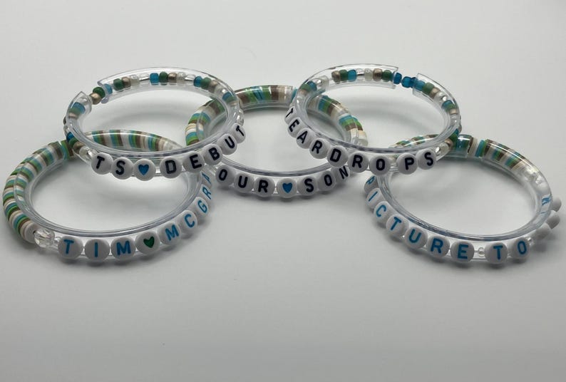 May include: Five clear bracelets with white letter beads spelling out phrases like "TS Debut" and "Picture to". The bracelets have a mix of white, blue, and green beads.