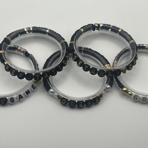 May include: Five clear bracelets with black and gold accents. Some bracelets have white beads spelling out words like "END GAME" and "GORGEOUS". The other bracelets have black beads spelling out words.
