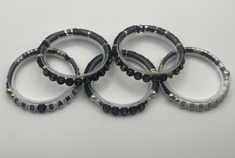 May include: Five clear bracelets with black and gold accents. Some bracelets have white beads spelling out words like "END GAME" and "GORGEOUS". The other bracelets have black beads spelling out words.
