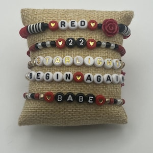 May include: Six beaded bracelets with white, red, black, and gold beads. The bracelets spell out "RED", "22", "STARLIGHT", "BEGIN AGAIN", and "BABE". The bracelets are displayed on a burlap pillow.