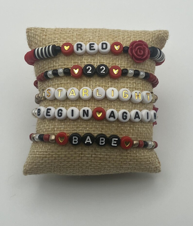 May include: Six beaded bracelets with white, red, black, and gold beads. The bracelets spell out "RED", "22", "STARLIGHT", "BEGIN AGAIN", and "BABE". The bracelets are displayed on a burlap pillow.