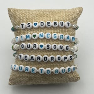 May include: Four beaded bracelets with white letter beads spelling out "TS DEBUT", "TIM MCGRAW", "OUR SONG", "TEARDROPS", and "PICTURE TO BURN". Each bracelet has heart-shaped beads and is displayed on a tan burlap pillow.