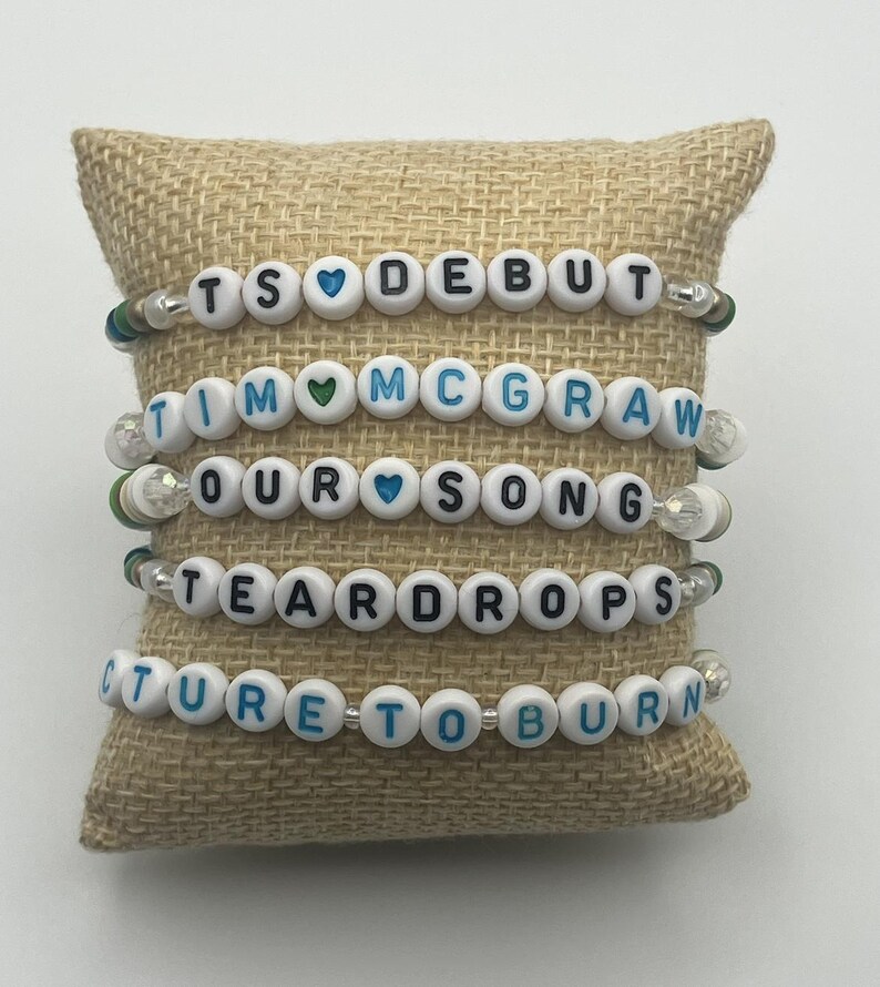 May include: Four beaded bracelets with white letter beads spelling out "TS DEBUT", "TIM MCGRAW", "OUR SONG", "TEARDROPS", and "PICTURE TO BURN". Each bracelet has heart-shaped beads and is displayed on a tan burlap pillow.