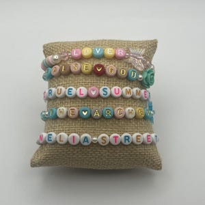 May include: Five beaded bracelets with colourful letter beads spelling out words like "LOVER", "FALSE GOD", and "CRUEL SUMMER". The bracelets are displayed on a small, textured, tan cushion.