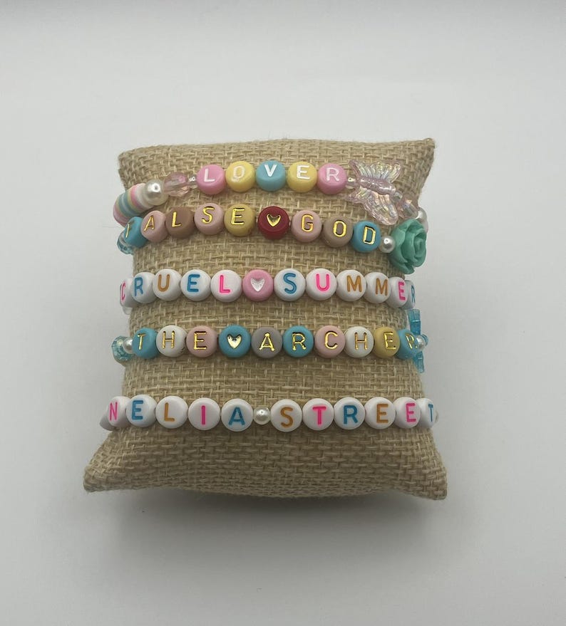 May include: Five beaded bracelets with colourful letter beads spelling out words like "LOVER", "FALSE GOD", and "CRUEL SUMMER". The bracelets are displayed on a small, textured, tan cushion.