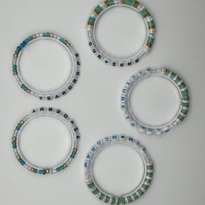 May include: Five clear bracelets with colorful beads and text. The bracelets have white, blue, green, and brown beads. The text on the bracelets includes the words "TEARDROPS", "DEBUT", "OUR SONG", "PICTURE TO", and "MCGRAW".