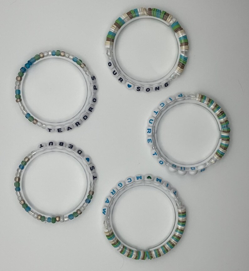 May include: Five clear bracelets with colorful beads and text. The bracelets have white, blue, green, and brown beads. The text on the bracelets includes the words "TEARDROPS", "DEBUT", "OUR SONG", "PICTURE TO", and "MCGRAW".