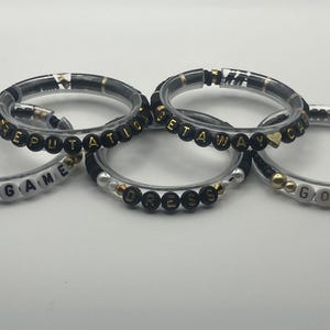 May include: Five clear bracelets with black and gold beads spelling out words like "END GAME", "REPUTATION", "GETAWAY", "DRESS", and "GORGEOUS". The bracelets have a mix of gold and black accents.