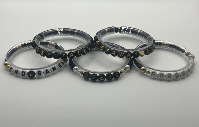 May include: Five clear bracelets with black and gold beads spelling out words like "END GAME", "REPUTATION", "GETAWAY", "DRESS", and "GORGEOUS". The bracelets have a mix of gold and black accents.
