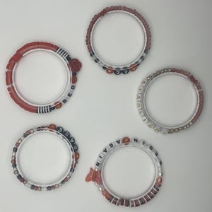 May include: Five clear bracelets with red, white, and blue beads. One bracelet has a rose and the word "RED". Another has the numbers "22". One bracelet says "BABE", another says "STARLIGHT", and the last says "AGAIN".