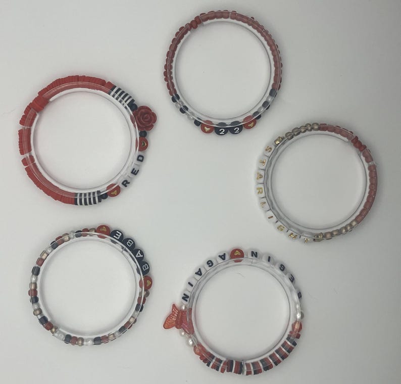 May include: Five clear bracelets with red, white, and blue beads. One bracelet has a rose and the word "RED". Another has the numbers "22". One bracelet says "BABE", another says "STARLIGHT", and the last says "AGAIN".