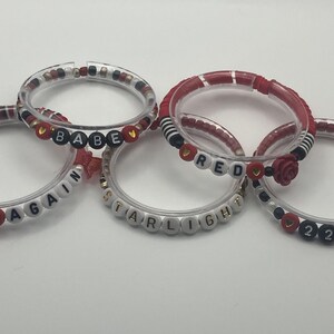 May include: Five clear bracelets with various bead designs. Some beads spell out words like "BEGIN AGAIN", "BABE", "STARLIGHT", and "RED". Other beads are red, black, and white, with a rose accent on one.