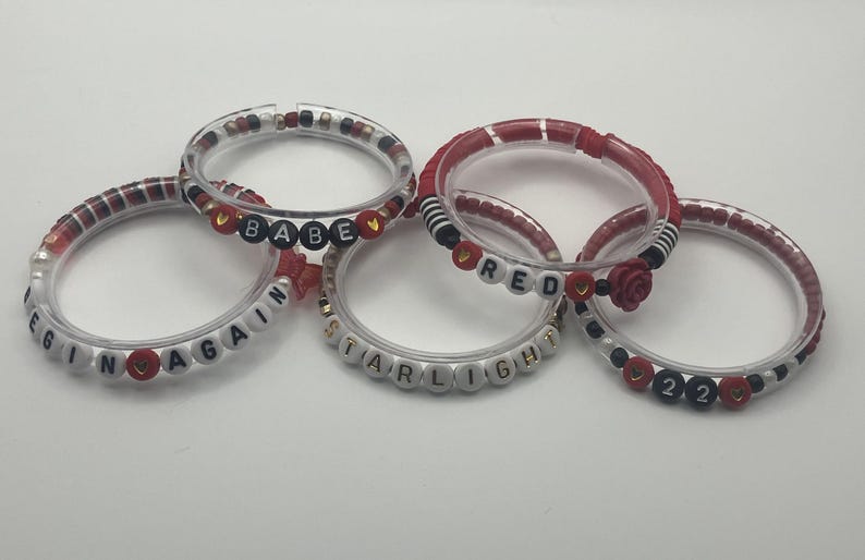 May include: Five clear bracelets with various bead designs. Some beads spell out words like "BEGIN AGAIN", "BABE", "STARLIGHT", and "RED". Other beads are red, black, and white, with a rose accent on one.