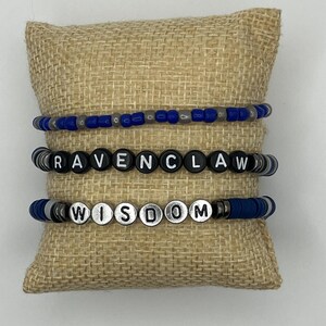 Könnte beinhalten: Drei Armbänder mit blauen, schwarzen und silbernen Perlen. Die Armbänder buchstabieren die Wörter "RAVENCLAW" und "WISDOM" mit Buchstabenperlen. Die Armbänder werden auf einem Jutekissen präsentiert.