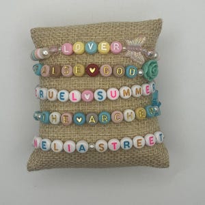 May include: Five beaded bracelets with colourful letter beads spelling out "LOVER", "FALSE GOD", "CRUEL SUMMER", "THE ARCHER", and "NELIA STREET". The bracelets are displayed on a burlap pillow.