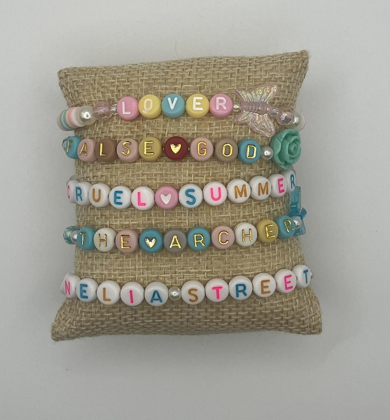 May include: Five beaded bracelets with colourful letter beads spelling out "LOVER", "FALSE GOD", "CRUEL SUMMER", "THE ARCHER", and "NELIA STREET". The bracelets are displayed on a burlap pillow.
