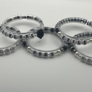 K&ouml;nnte beinhalten: F&uuml;nf transparente Armb&auml;nder mit wei&szlig;en Buchstabenperlen, die W&ouml;rter wie "CARDIGAN", "FOLKLORE", "HOAX", "EXILE" und "BETTY" buchstabieren. Die Armb&auml;nder haben schwarze und wei&szlig;e Perlen und kleine Perlen.