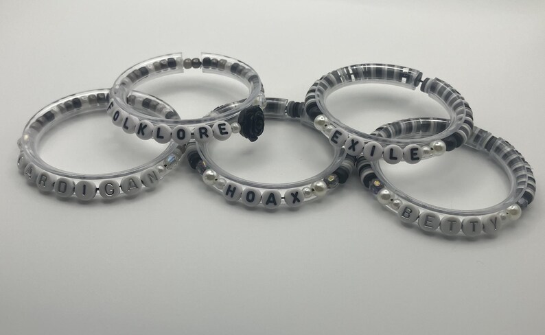 K&ouml;nnte beinhalten: F&uuml;nf transparente Armb&auml;nder mit wei&szlig;en Buchstabenperlen, die W&ouml;rter wie "CARDIGAN", "FOLKLORE", "HOAX", "EXILE" und "BETTY" buchstabieren. Die Armb&auml;nder haben schwarze und wei&szlig;e Perlen und kleine Perlen.
