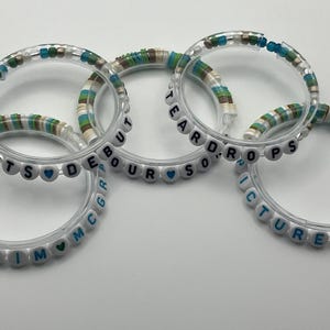 May include: Five clear bracelets with white letter beads spelling out words and names. The bracelets have accents of blue, green, and tan beads. The words include "TEARDROPS", "PICTURE TO", "DEBOUR", and "TIM MCGRAW".