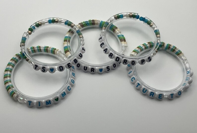 May include: Five clear bracelets with white letter beads spelling out words and names. The bracelets have accents of blue, green, and tan beads. The words include "TEARDROPS", "PICTURE TO", "DEBOUR", and "TIM MCGRAW".