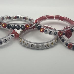 May include: Five clear bracelets with various designs. Some bracelets have red and black accents, while others feature white letter beads spelling out words like "BEGIN AGAIN", "BABE", "RED", "STARLIGHT", and "22".
