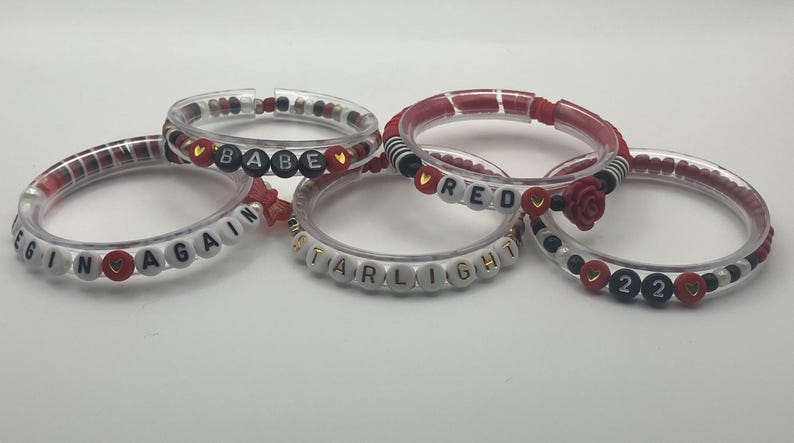 May include: Five clear bracelets with various designs. Some bracelets have red and black accents, while others feature white letter beads spelling out words like "BEGIN AGAIN", "BABE", "RED", "STARLIGHT", and "22".