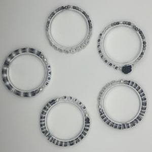 K&ouml;nnte beinhalten: F&uuml;nf transparente Armb&auml;nder mit verschiedenen Designs. Einige haben schwarz-wei&szlig;e Perlen, andere zeigen wei&szlig;e Buchstabenperlen mit W&ouml;rtern wie "HOAX", "EXILE" und "FOLKLORE". Ein Armband hat einen kleinen schwarzen Rosenanh&auml;nger.