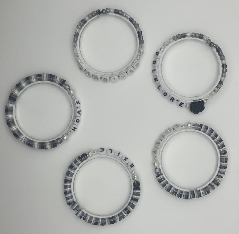 K&ouml;nnte beinhalten: F&uuml;nf transparente Armb&auml;nder mit verschiedenen Designs. Einige haben schwarz-wei&szlig;e Perlen, andere zeigen wei&szlig;e Buchstabenperlen mit W&ouml;rtern wie "HOAX", "EXILE" und "FOLKLORE". Ein Armband hat einen kleinen schwarzen Rosenanh&auml;nger.