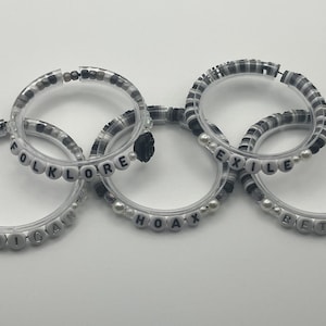 K&ouml;nnte beinhalten: F&uuml;nf transparente Armb&auml;nder mit schwarzen und wei&szlig;en Perlen und wei&szlig;en Buchstabenperlen mit den W&ouml;rtern "cardigan", "folklore", "hoax", "exile" und "betty". Die Armb&auml;nder sind mit einer schwarzen Blume verbunden.