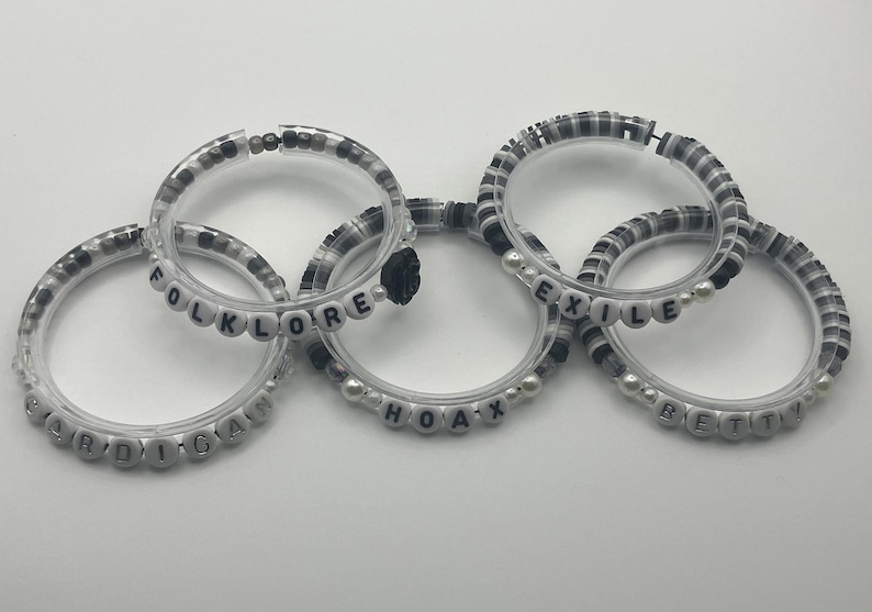 K&ouml;nnte beinhalten: F&uuml;nf transparente Armb&auml;nder mit schwarzen und wei&szlig;en Perlen und wei&szlig;en Buchstabenperlen mit den W&ouml;rtern "cardigan", "folklore", "hoax", "exile" und "betty". Die Armb&auml;nder sind mit einer schwarzen Blume verbunden.