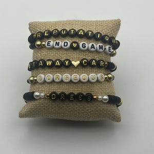 May include: Five beaded bracelets with black and gold letter beads spelling out "REPUTATION", "END GAME", "GETAWAY CAR", "GORGEOUS", and "DRESS". The bracelets are displayed on a burlap pillow.