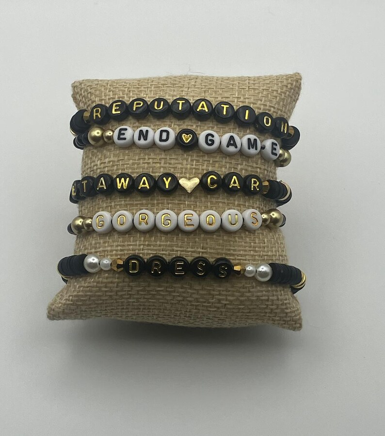 May include: Five beaded bracelets with black and gold letter beads spelling out "REPUTATION", "END GAME", "GETAWAY CAR", "GORGEOUS", and "DRESS". The bracelets are displayed on a burlap pillow.
