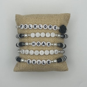 K&ouml;nnte beinhalten: F&uuml;nf Armb&auml;nder mit Perlen mit wei&szlig;en Buchstaben, die "FOLKLORE", "BETTY", "HOAX", "CARDIGAN" und "EXILE" buchstabieren. Die Armb&auml;nder haben schwarze und wei&szlig;e Perlen und werden auf einem Jutekissen pr&auml;sentiert.