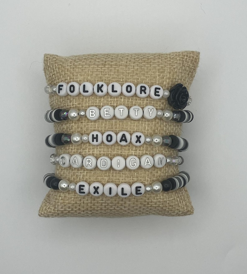K&ouml;nnte beinhalten: F&uuml;nf Armb&auml;nder mit Perlen mit wei&szlig;en Buchstaben, die "FOLKLORE", "BETTY", "HOAX", "CARDIGAN" und "EXILE" buchstabieren. Die Armb&auml;nder haben schwarze und wei&szlig;e Perlen und werden auf einem Jutekissen pr&auml;sentiert.