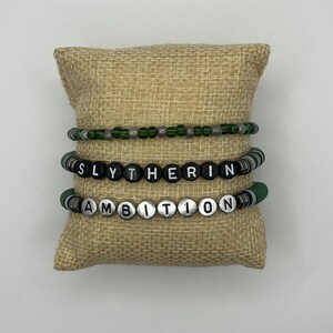 Könnte beinhalten: Drei Perlenarmbänder in Grün, Schwarz und Silber, präsentiert auf einem Jutekissen. Das mittlere Armband schreibt "SLYTHERIN" in schwarzen und weißen Perlen. Das untere Armband schreibt "AMBITION" in silbernen und schwarzen Perlen.