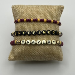 以下が含まれることがあります： 赤、黄、黒のビーズブレスレット3本。「GRYFFINDOR」と「BRAVERY」の文字が金色の文字で書かれています。ブレスレットはジュートの枕の上に展示されています。