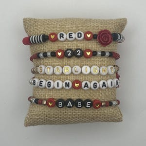 May include: Five beaded bracelets with various word combinations, including "RED", "22", "STARLIGHT", "BEGIN AGAIN", and "BABE". The bracelets feature red, black, white, and gold beads, with heart accents and a rose detail.