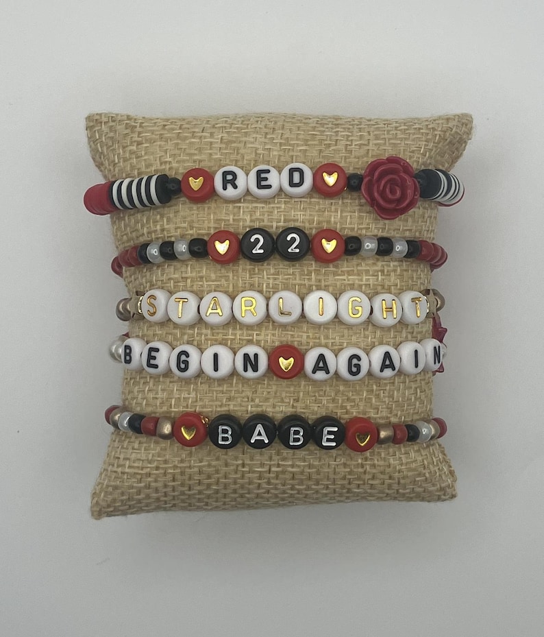 May include: Five beaded bracelets with various word combinations, including "RED", "22", "STARLIGHT", "BEGIN AGAIN", and "BABE". The bracelets feature red, black, white, and gold beads, with heart accents and a rose detail.